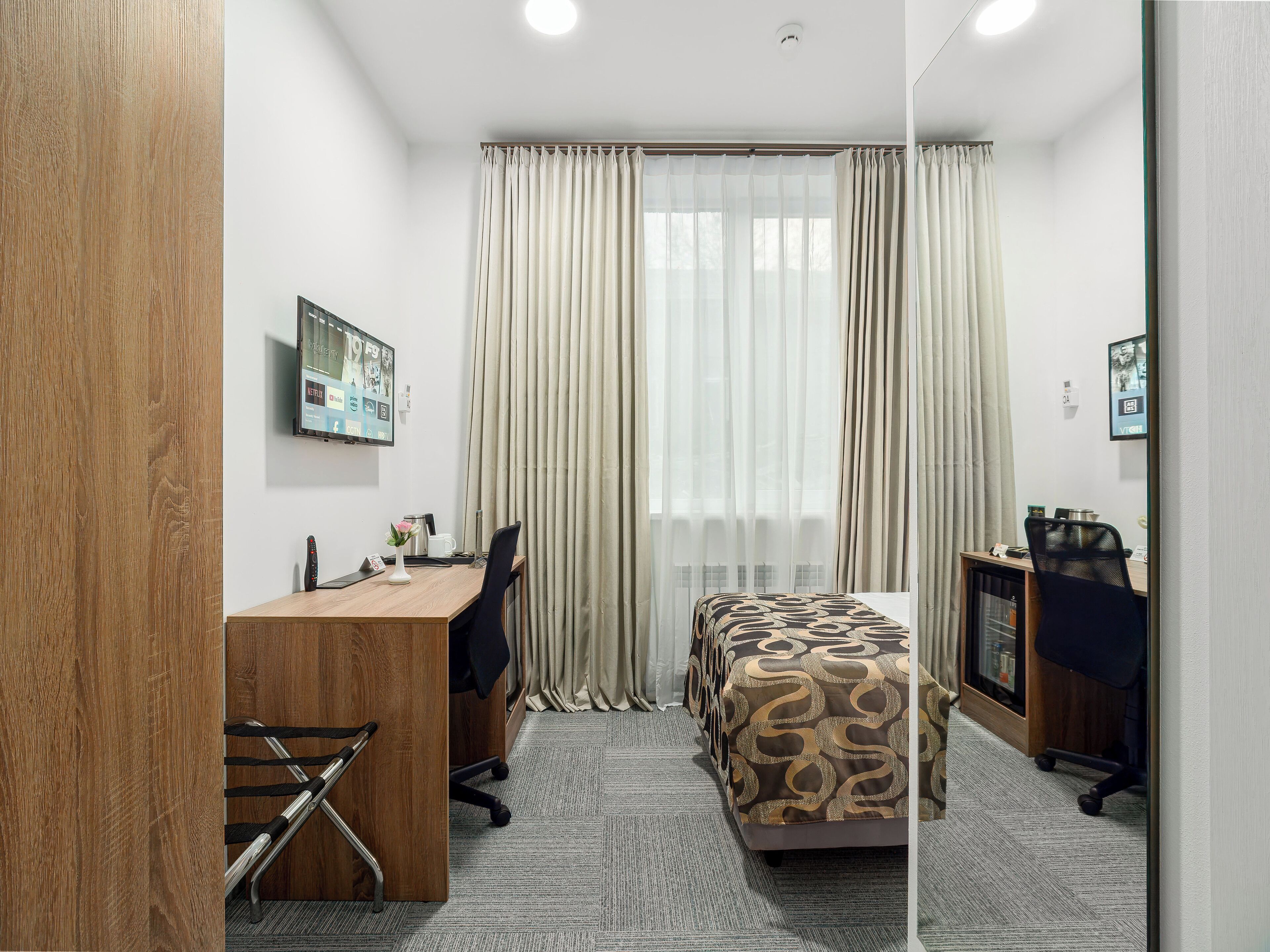 A hotel room with a wooden desk, a bed with patterned bedding, a TV mounted on the wall, and a window with curtains.