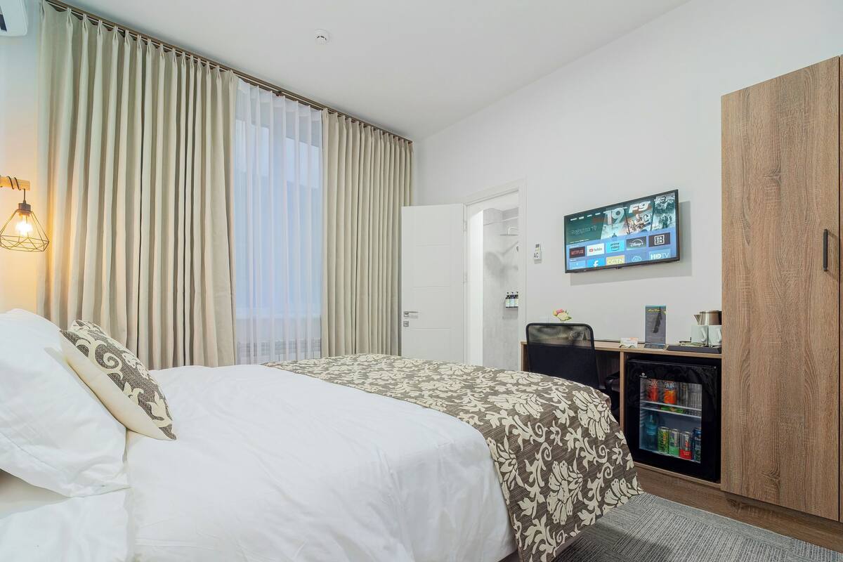 A hotel room with a bed, a desk with a computer, a TV, and a minibar.