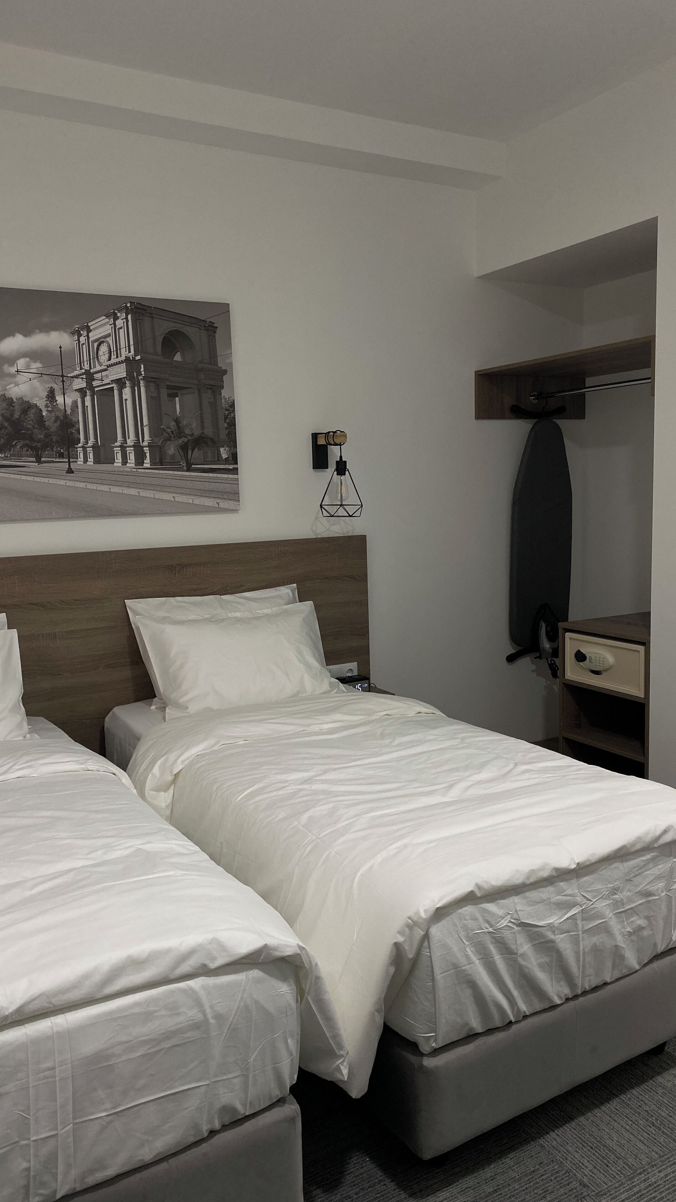A hotel room with two beds, a wooden headboard, a wall-mounted clock, a painting, a desk with a mirror, and a hanging lamp.