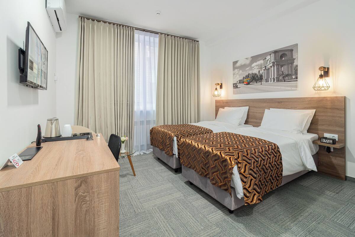 A hotel room with two beds, a desk, a TV, and a window with curtains.