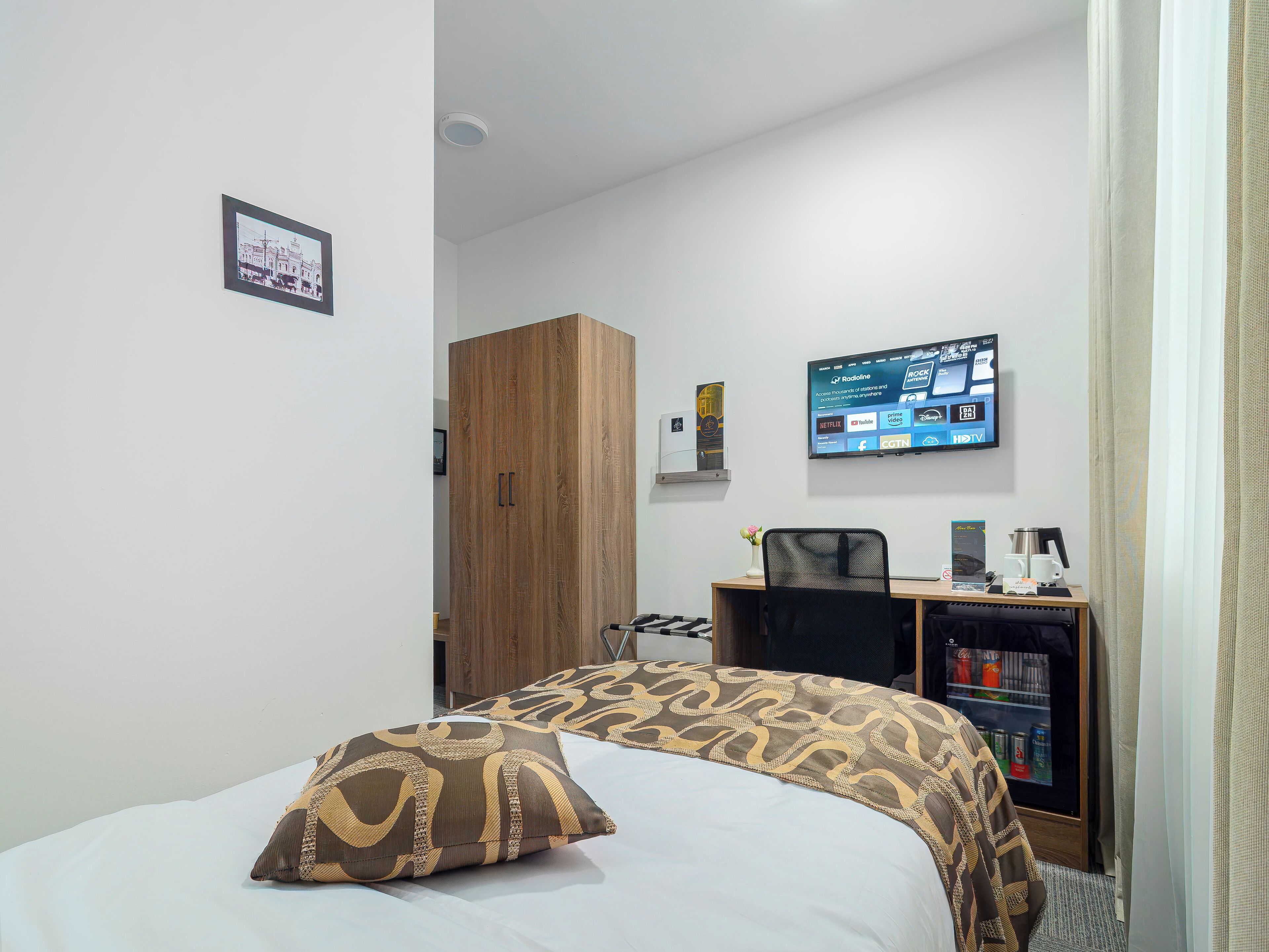 A hotel room with a bed, a desk with a computer, a TV mounted on the wall, and a wardrobe.