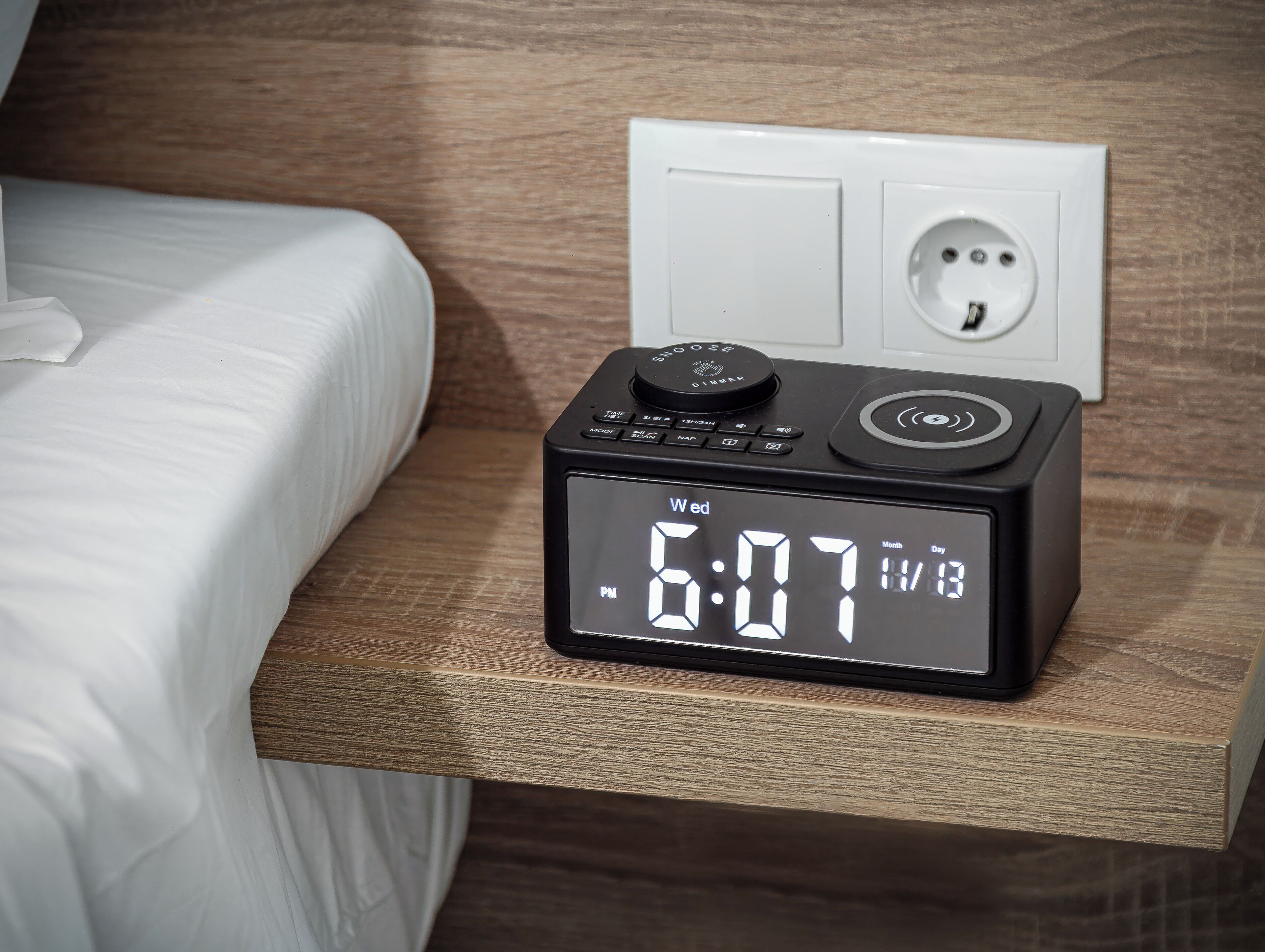 Une horloge numérique posée sur une table de chevet affiche l’heure : 18 h 07. On aperçoit en arrière-plan une prise de courant blanche et un lit recouvert de draps blancs.
