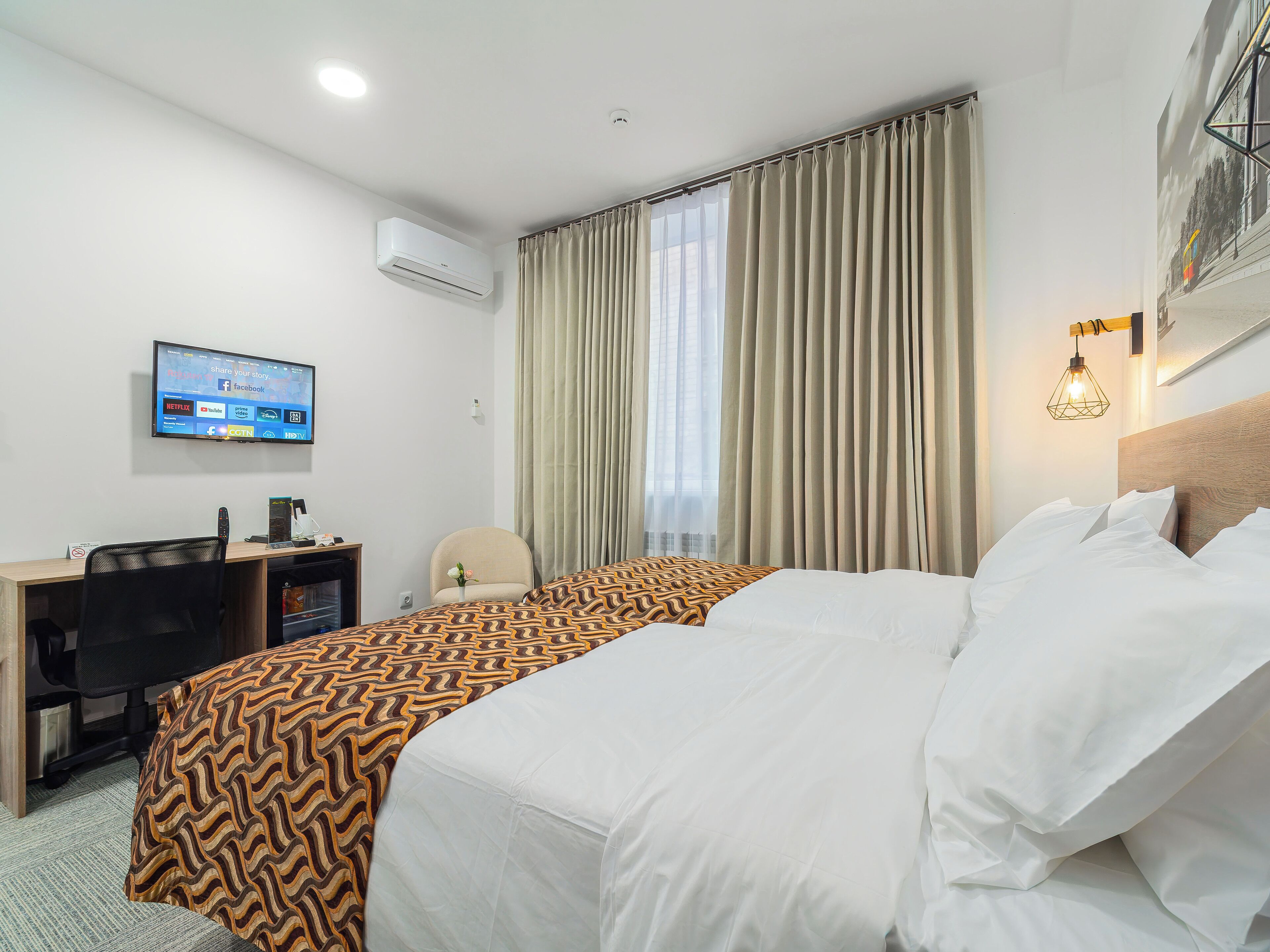 A hotel room with a bed, a desk with a computer, a TV mounted on the wall, and a window with curtains.