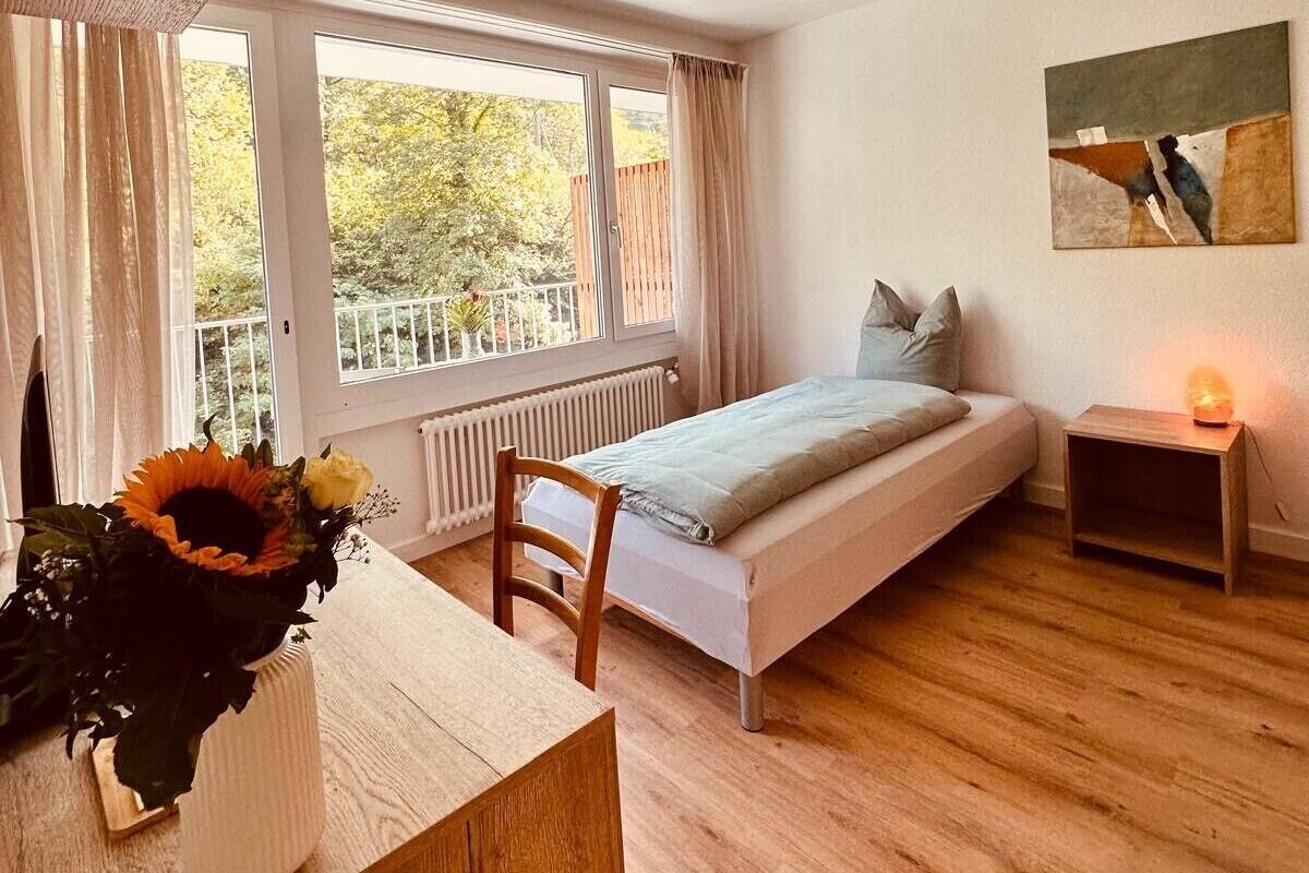 A bedroom with a bed, a bedside table, a chair, a window with curtains, and a painting on the wall.