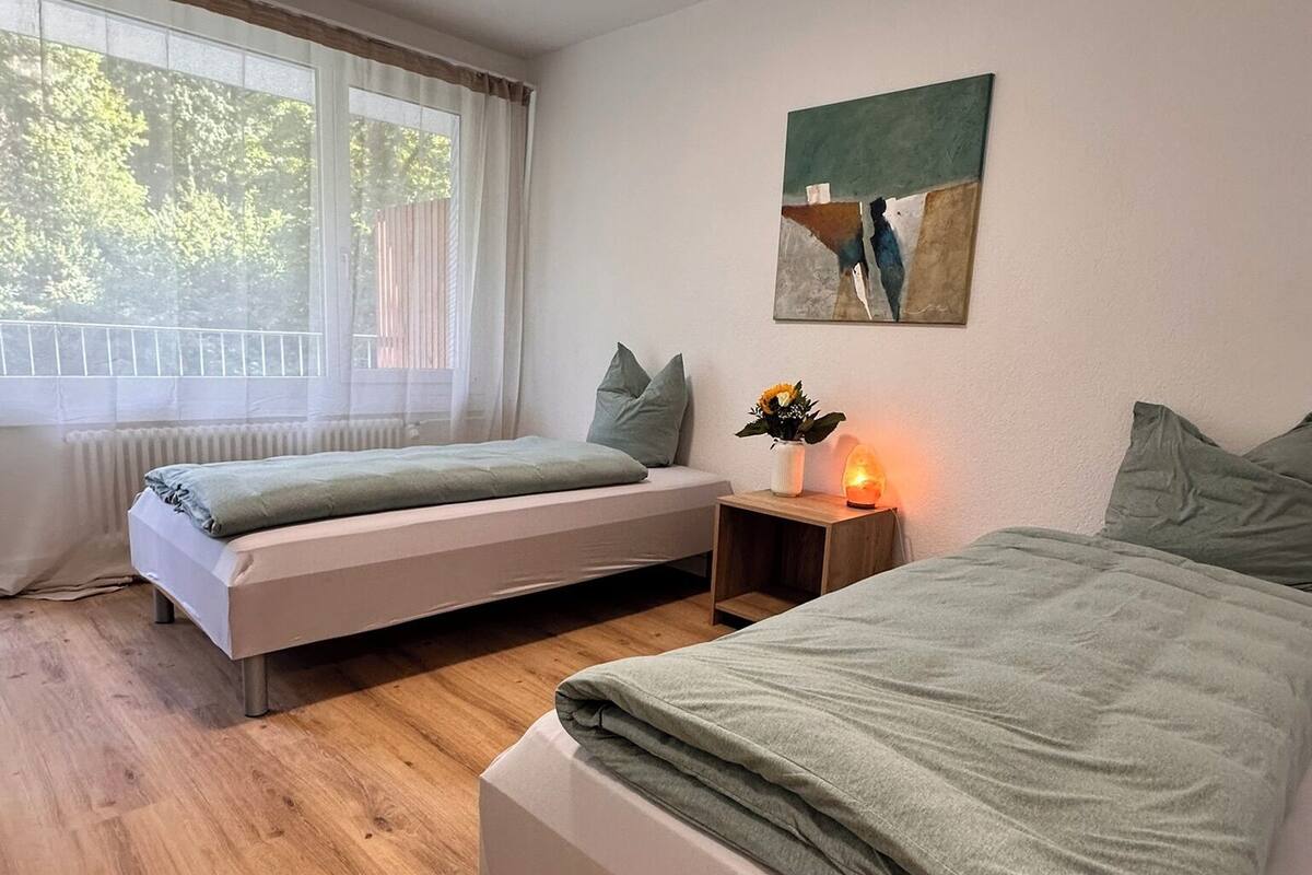A bedroom with two single beds, a wooden floor, a window with curtains, a bedside table with a lamp, and a painting on the wall.