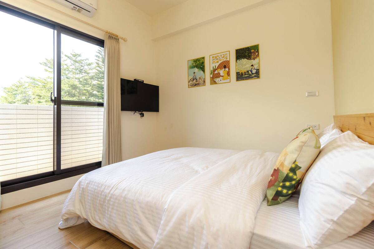 A bedroom with a large window, a bed with white bedding, and three wall-mounted paintings.