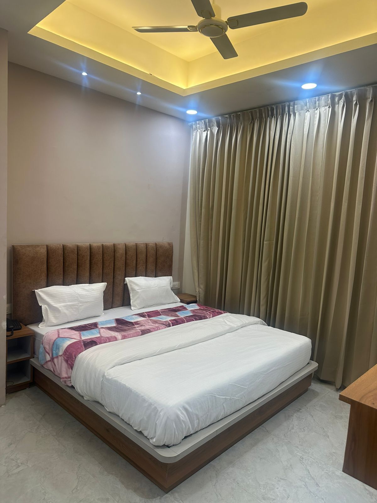 A modern hotel room with a large bed, a ceiling fan, and a view of the outdoors through curtains.