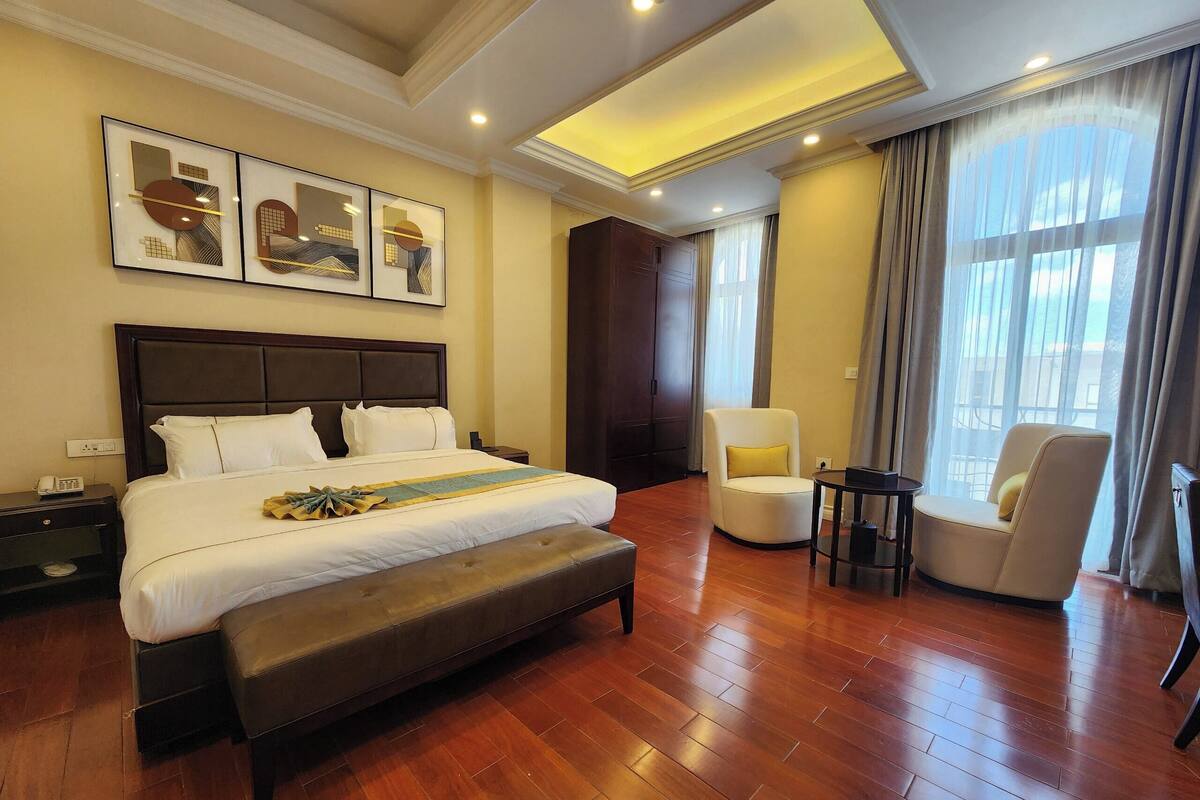 Une chambre d’hôtel avec un grand lit, une table de chevet avec un téléphone, une chaise, une petite table et une fenêtre avec des rideaux.