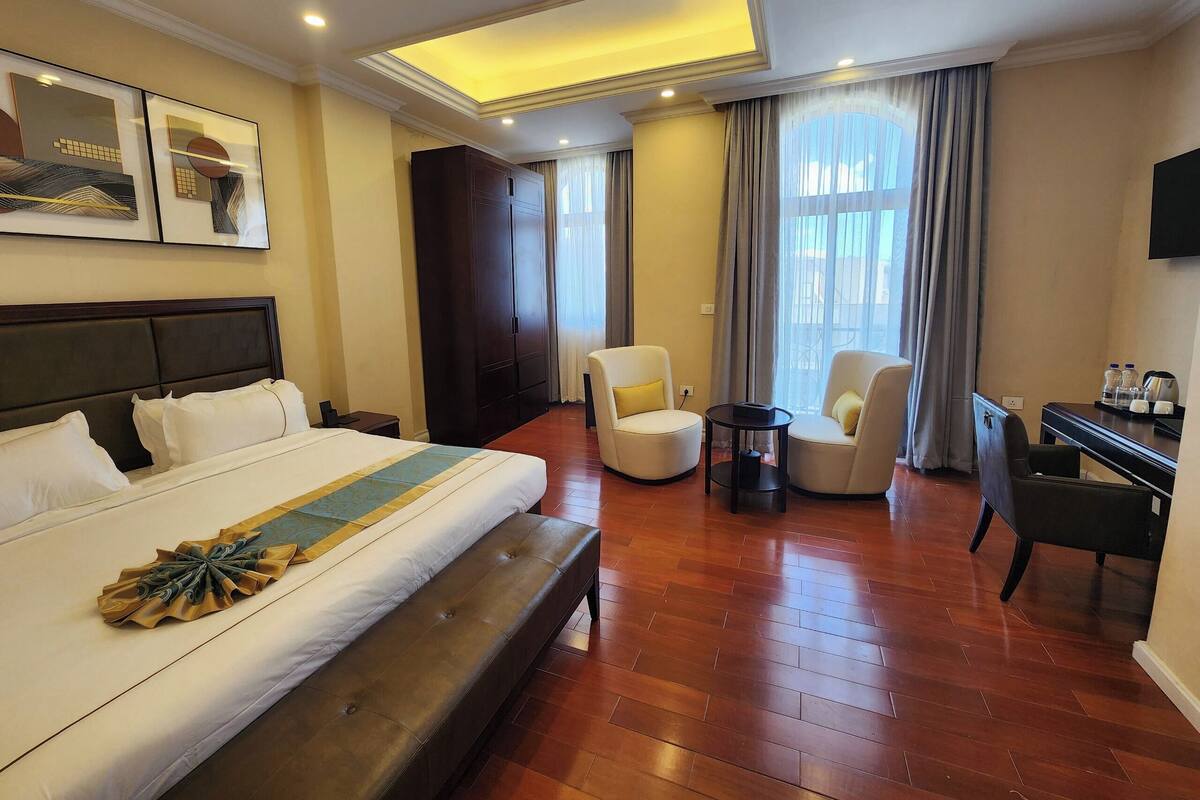 Une chambre d’hôtel avec un grand lit, deux fauteuils, un bureau, un téléviseur à écran plat et une grande fenêtre avec des rideaux.