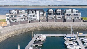 Une propriété située au bord de l’eau, dotée d’une architecture moderne, d’un port de plaisance avec plusieurs bateaux, et offrant une vue sur la mer et les éoliennes au loin.