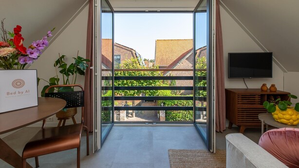 A modern living room with a large sliding glass door leading to a balcony, a flat-screen TV, and a dining area with a table and chairs. - Appartement Bed by Al - Hoofdstraat 16 a Kortgene (Kortgene)