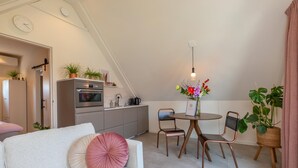 A cozy living room with a white sofa, a dining table with two chairs, a kitchen area, and a large window. - Appartement Bed by Al - Hoofdstraat 16 a Kortgene (Kortgene)