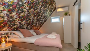 A bedroom with a bed, pillows, a bedside table with a lamp, a floral wall, a small window, and a door. - Appartement Bed by Al - Hoofdstraat 16 a Kortgene (Kortgene)