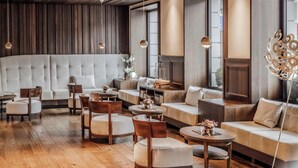 A modern interior with a sofa, chairs, and tables arranged around a central area with a wooden ceiling and large windows. - HOTIDAY Room Collection - Ortisei (Ortisei)