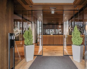 A hotel reception area with a wooden reception desk, glass doors, and potted plants. - HOTIDAY Room Collection - Ortisei (Ortisei)