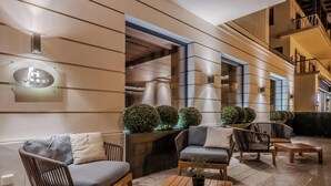 A modern outdoor seating area with cushioned chairs and a wooden table, a glass sign with a hotel logo, and a well-lit entrance. - HOTIDAY Room Collection - Ortisei (Ortisei)