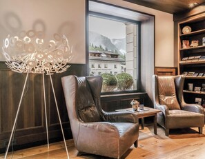 A modern room with leather chairs, a wooden floor, a large window with a view of buildings and mountains, and a bookshelf with books and decorative items. - HOTIDAY Room Collection - Ortisei (Ortisei)
