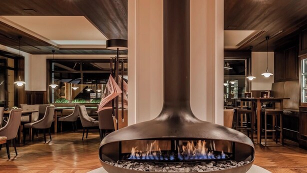 A modern interior with a central fire feature, wooden flooring, and a dining area with tables and chairs. - HOTIDAY Room Collection - Ortisei (Ortisei)