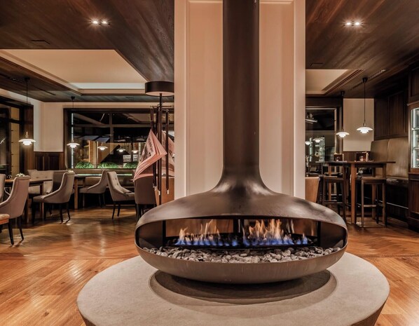 A modern interior with a central fire feature, wooden flooring, and a dining area with tables and chairs. - HOTIDAY Room Collection - Ortisei (Ortisei)