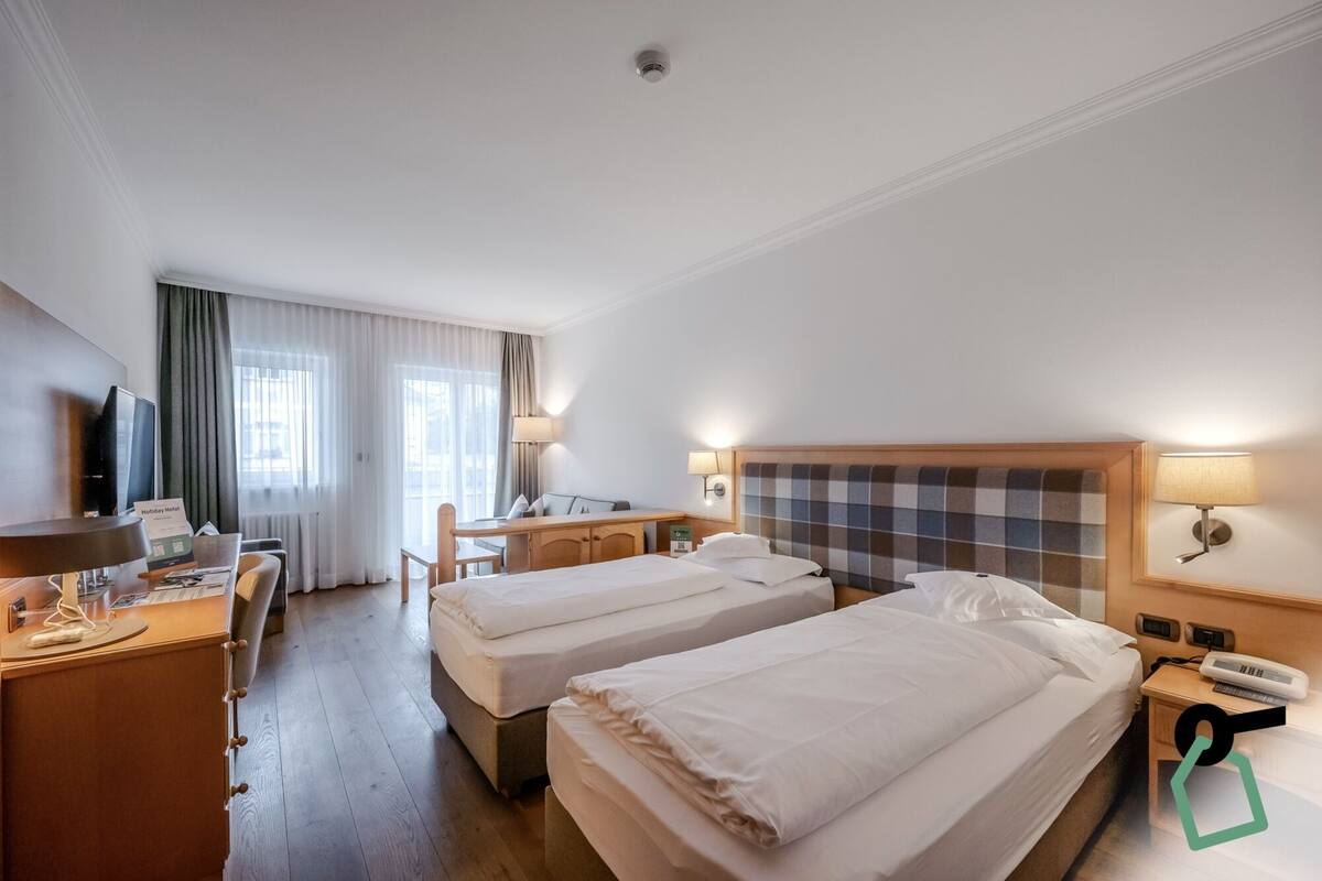 A hotel room with two beds, a desk, a chair, a TV, and a lamp.