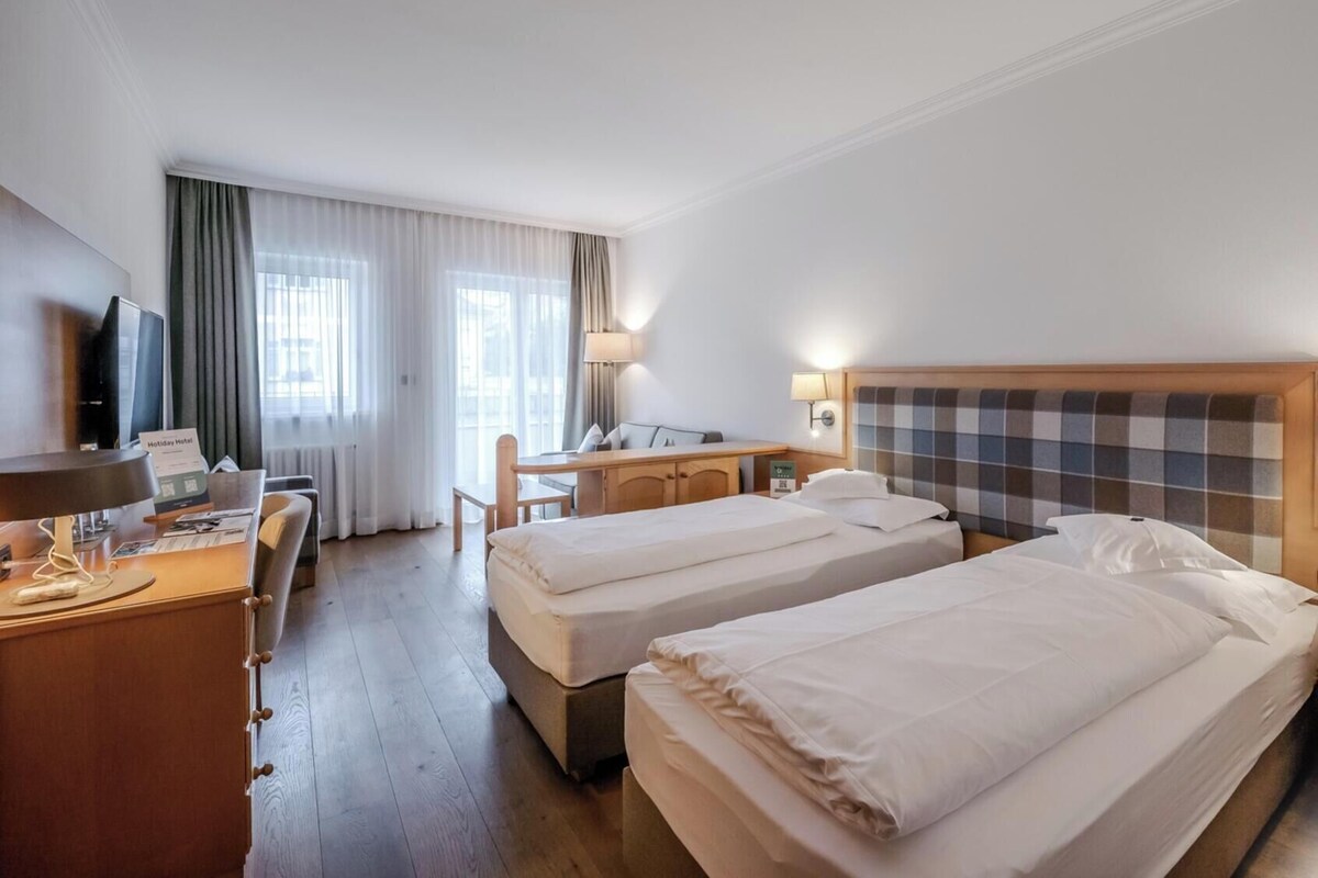 A hotel room with two beds, a desk, a chair, a TV, and a lamp.