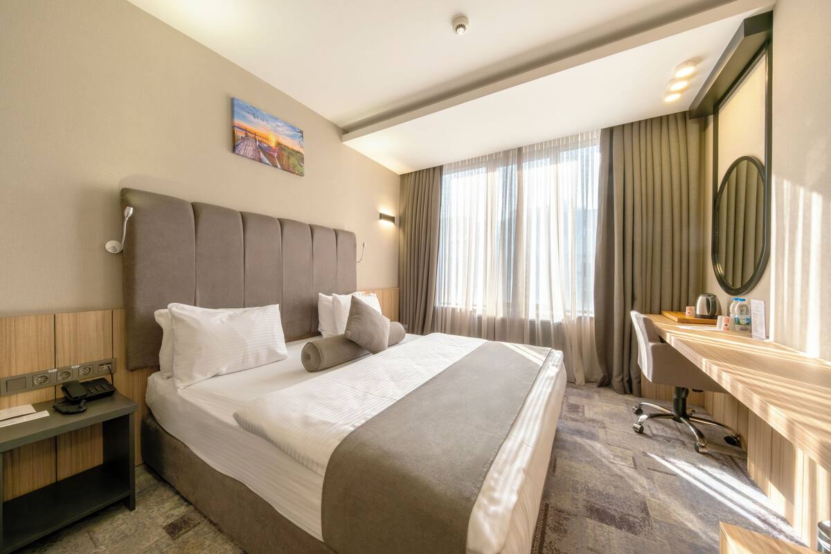 A hotel room with a large bed, a desk, a chair, a telephone, and a painting on the wall.