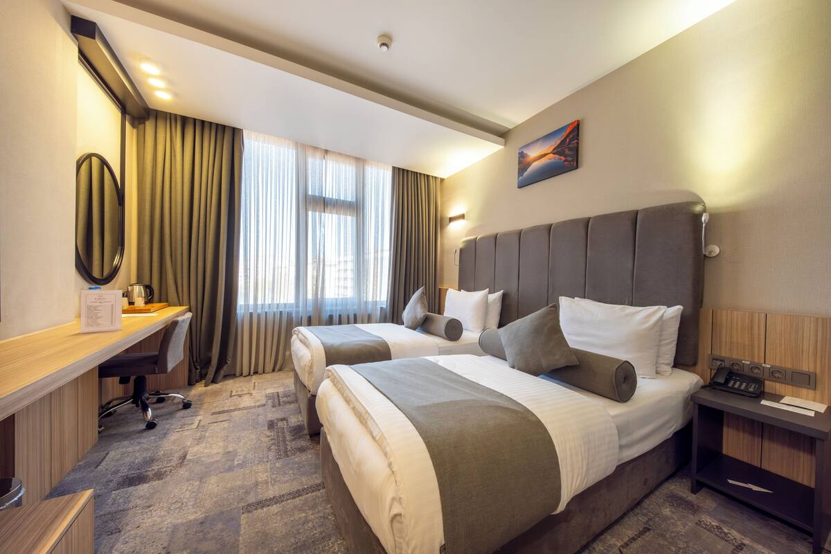 A hotel room with two beds, a desk, a chair, a nightstand, and a painting on the wall.