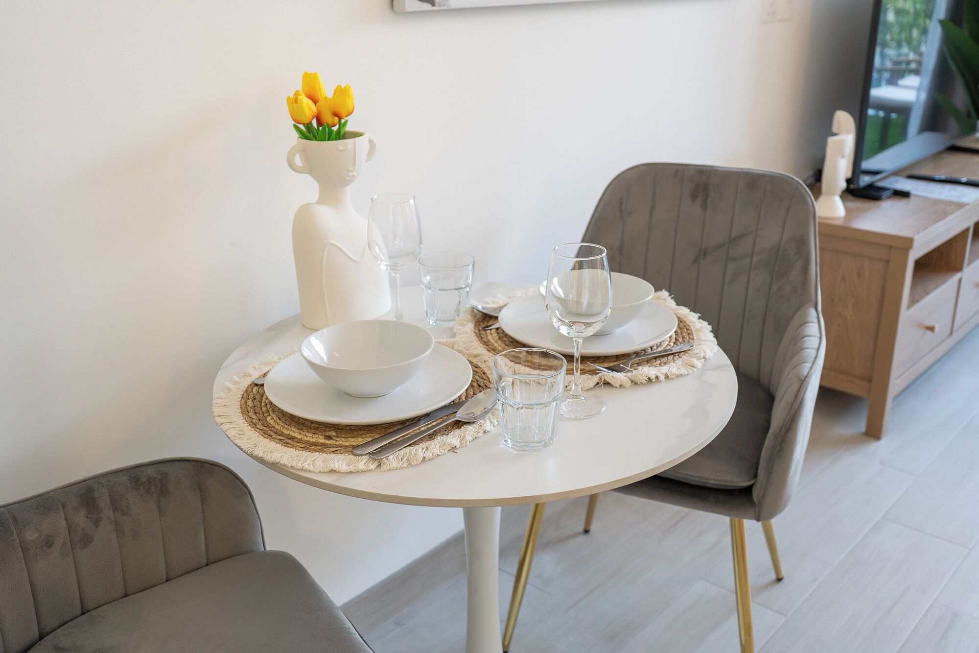 A dining table set with white plates, bowls, and glasses, a vase with yellow flowers, and a white pitcher.