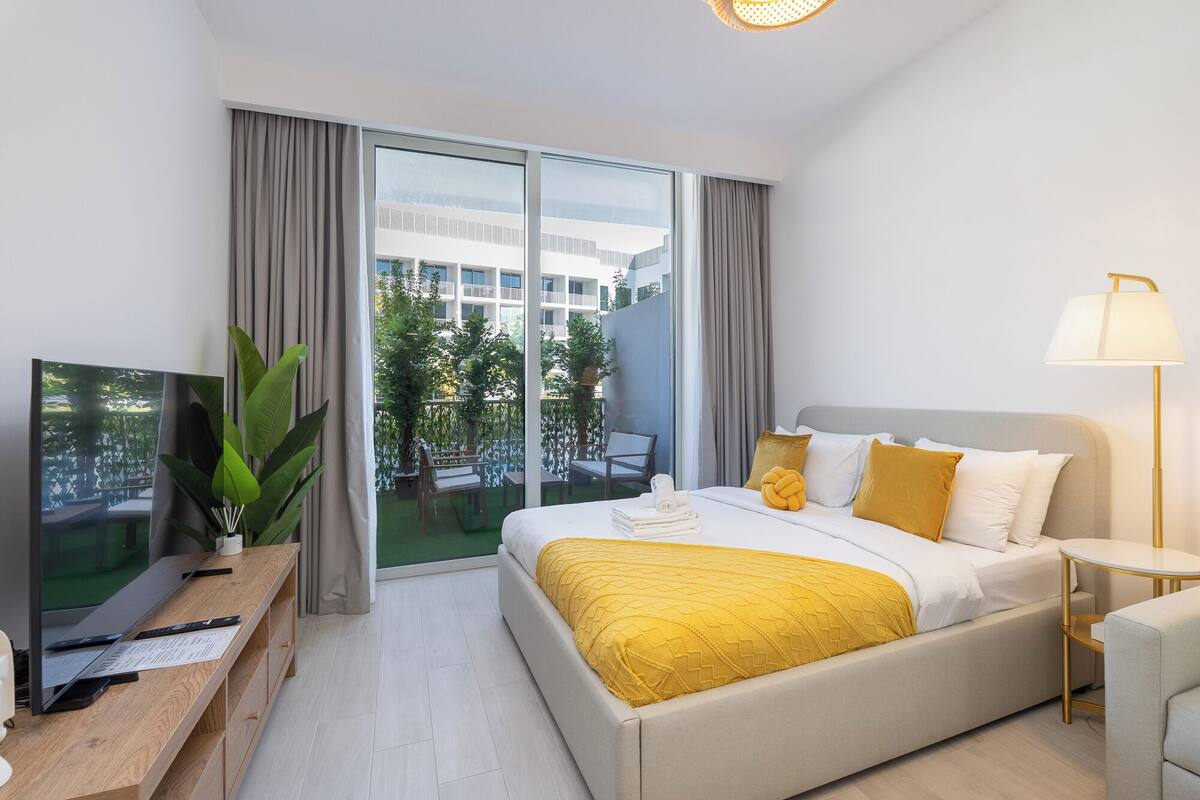 Une chambre d’hôtel avec un grand lit, un bureau avec un ordinateur, une télévision et un balcon offrant une vue sur les bâtiments et la verdure.