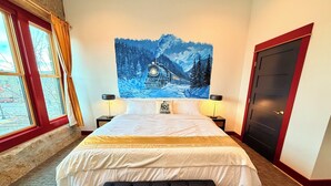 Hotelkamer met een groot bed, een bankje, twee nachtkastjes met lampen, een schilderij van een besneeuwd berglandschap, een raam met gordijnen en een deur.