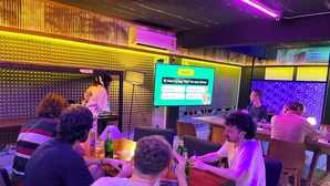 Een groep mensen zit aan tafels in een ruimte, terwijl een groot scherm een quiz toont.