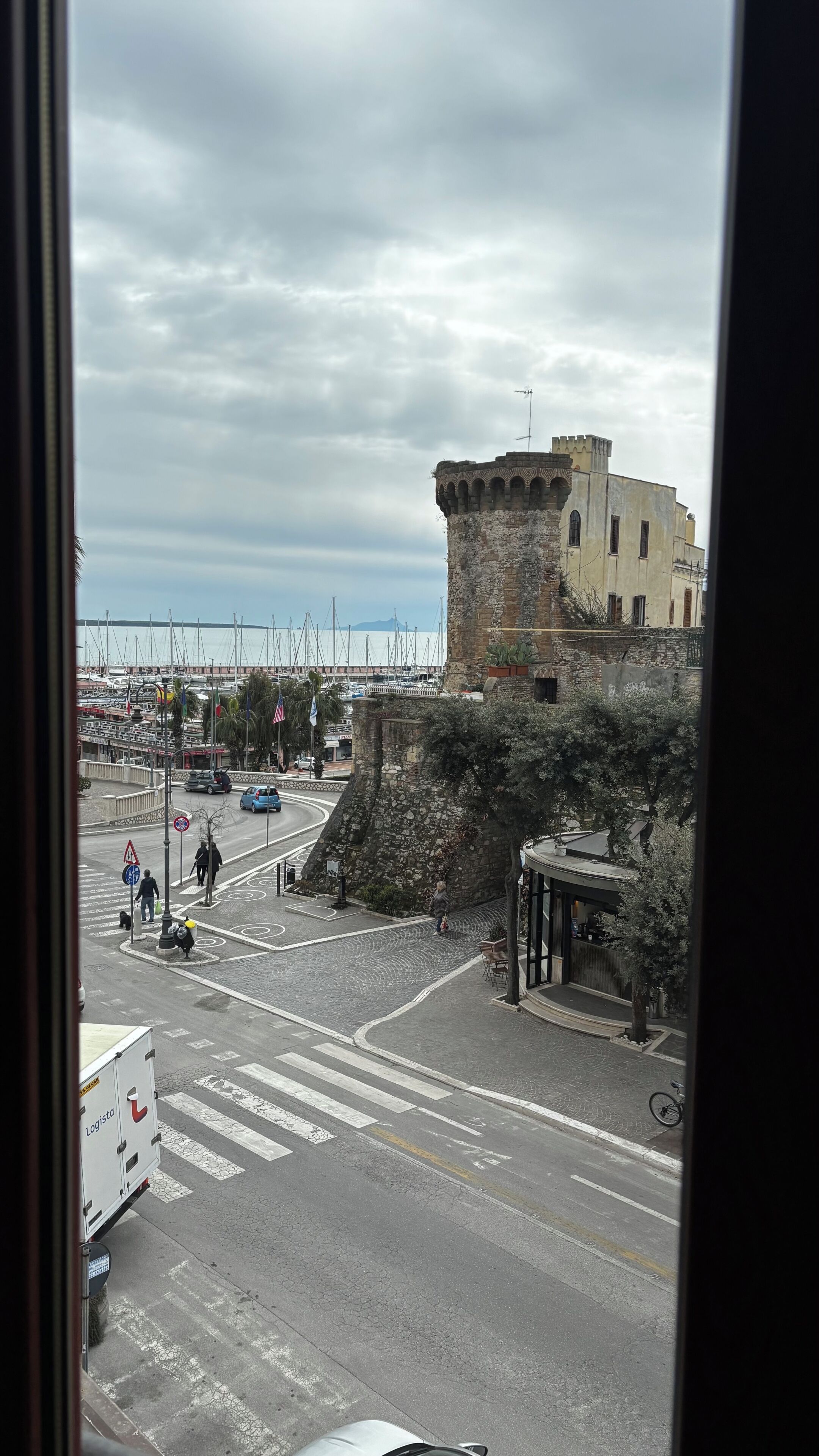 Une vue d’un bâtiment historique doté d’une tour, d’un port de plaisance avec des bateaux et d’une rue où l’on peut voir des piétons et des véhicules.