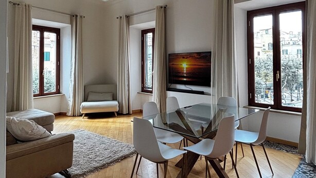 Un salón espacioso con techo de madera, televisor de pantalla plana, mesa de vidrio con sillas, sofá y ventanas grandes con cortinas.