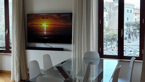 Un comedor moderno con una mesa de vidrio, sillas blancas, un televisor de pantalla plana y ventanas amplias con cortinas.