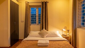 Una habitación de hotel con cama, cortinas, ventana, mesita de noche con lámpara y puerta.