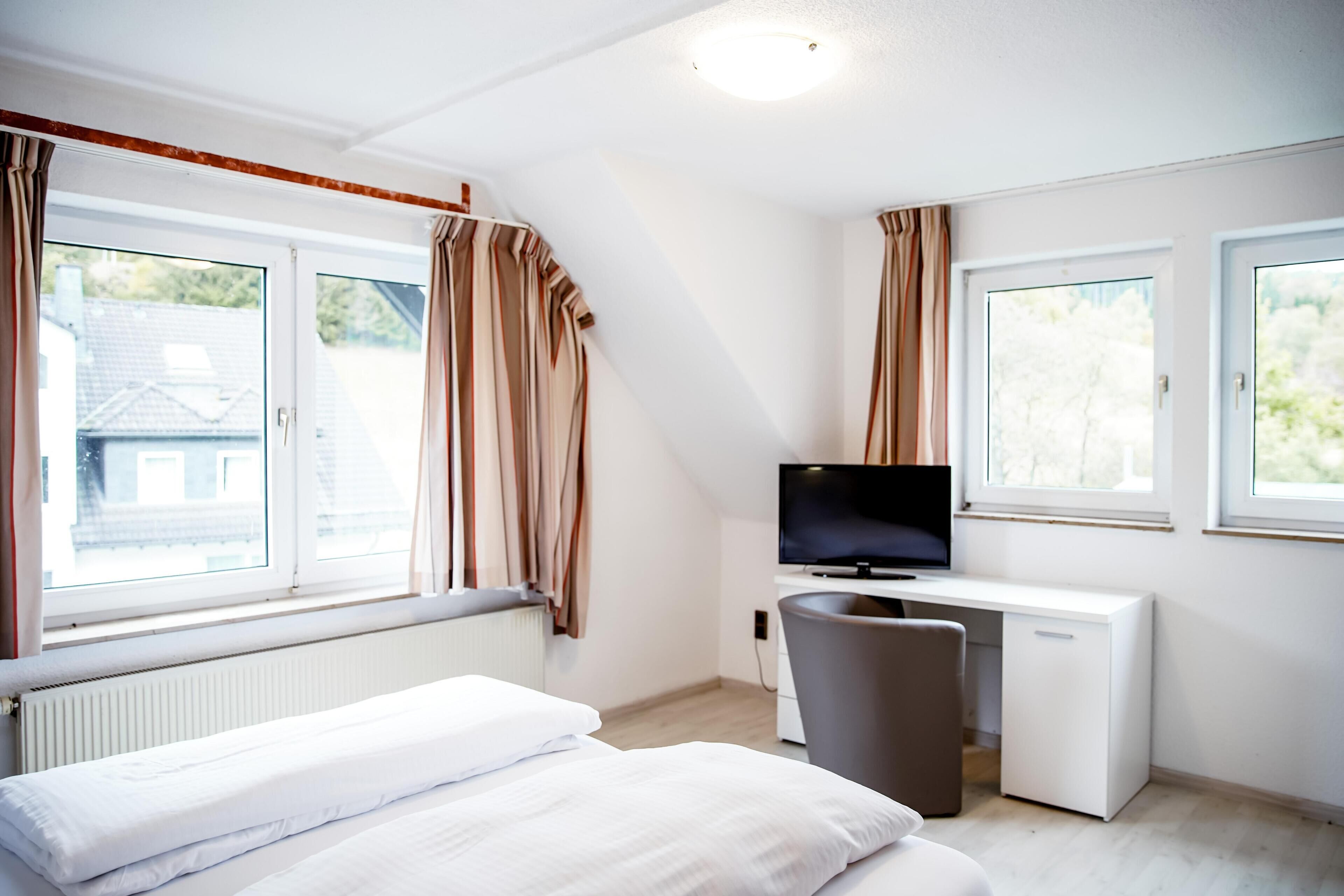 Una habitación de hotel moderna con una cama, un escritorio con televisor y ventanas amplias con cortinas.