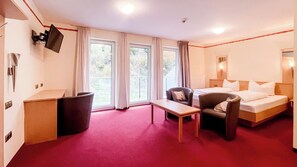 A hotel room with a bed, a desk, a chair, a TV mounted on the wall, and large windows with curtains.