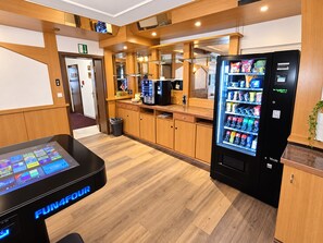 A hotel lobby with wooden flooring, a vending machine, a digital kiosk, and a trash bin. - Comfortable and versatile room -perfect for relaxation, work and exchange (Winterberg)