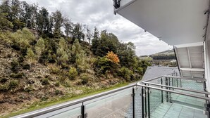 Un balcon doté d’une balustrade en verre, donnant sur une colline boisée et offrant une vue lointaine sur une maison.