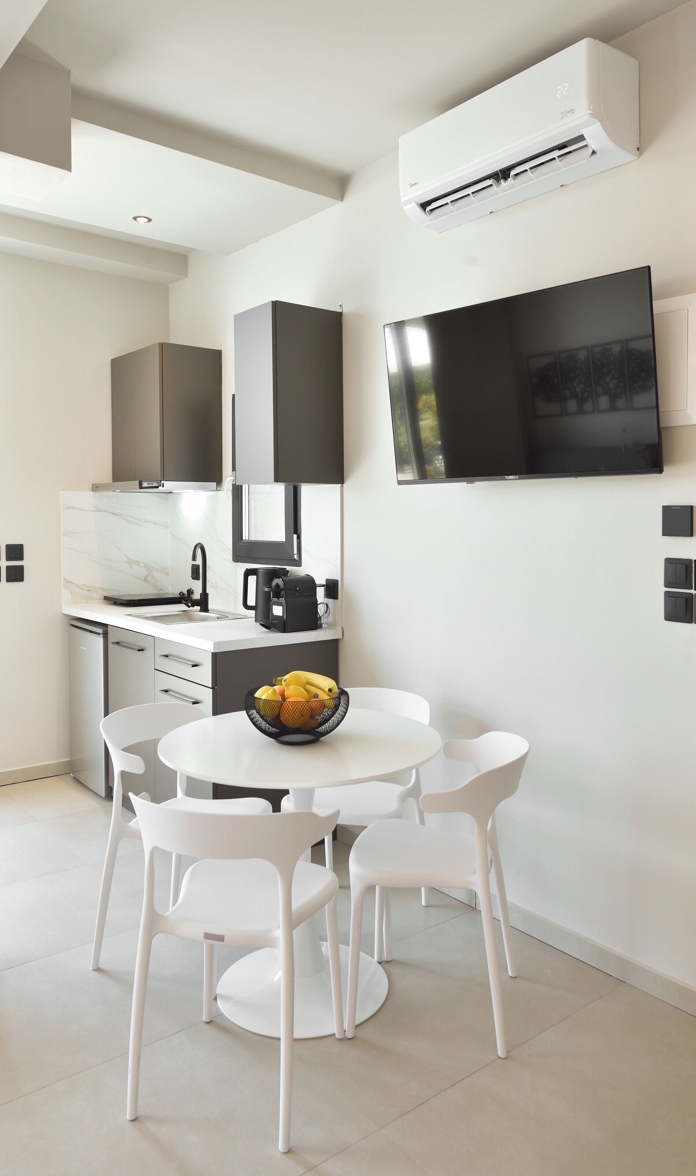 A modern kitchen with white cabinetry, a built-in oven, and a coffee machine. There is a dining area with a white table and chairs, and a bowl of fruit on the table.