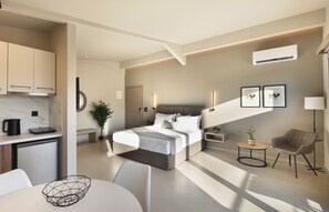 A modern hotel room with a bed, bedside tables, a desk, a chair, a small kitchen area, and an air conditioning unit.