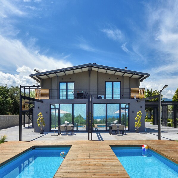 A modern house with a large glass front, a pool, and outdoor furniture.