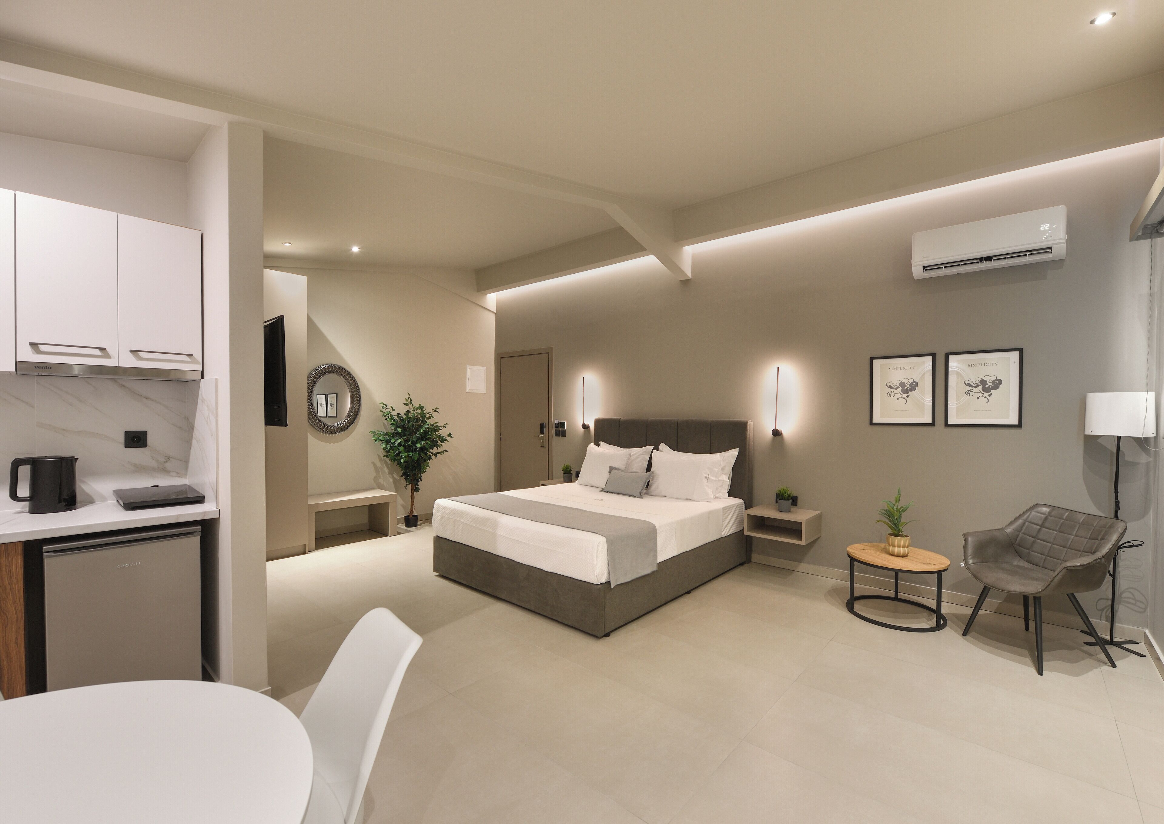 A modern hotel room with a large bed, bedside tables, a desk with a chair, a small round table with a chair, and a potted plant.