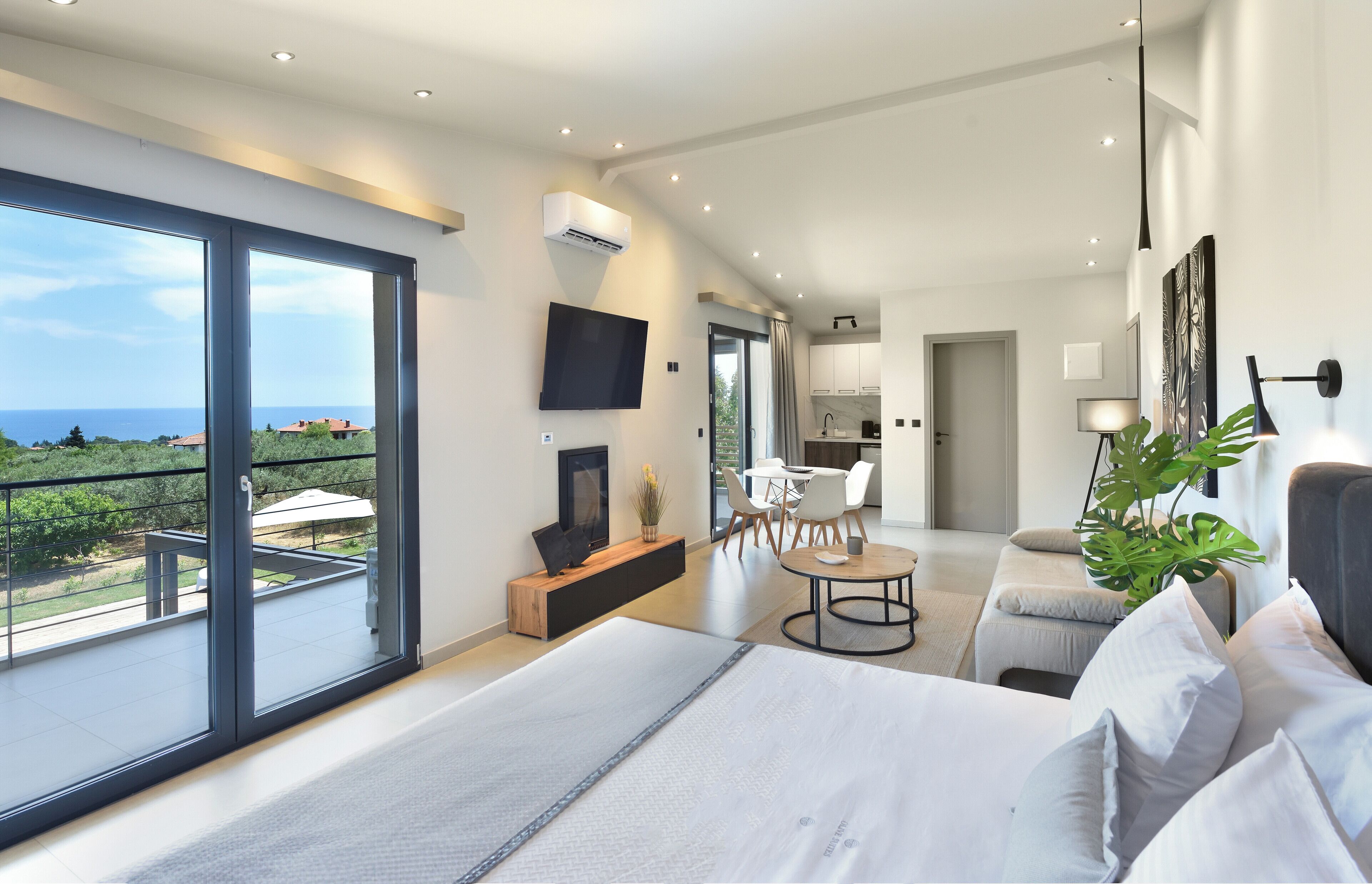 A modern bedroom with a large bed, a TV, a balcony with a view, and a dining area with chairs.