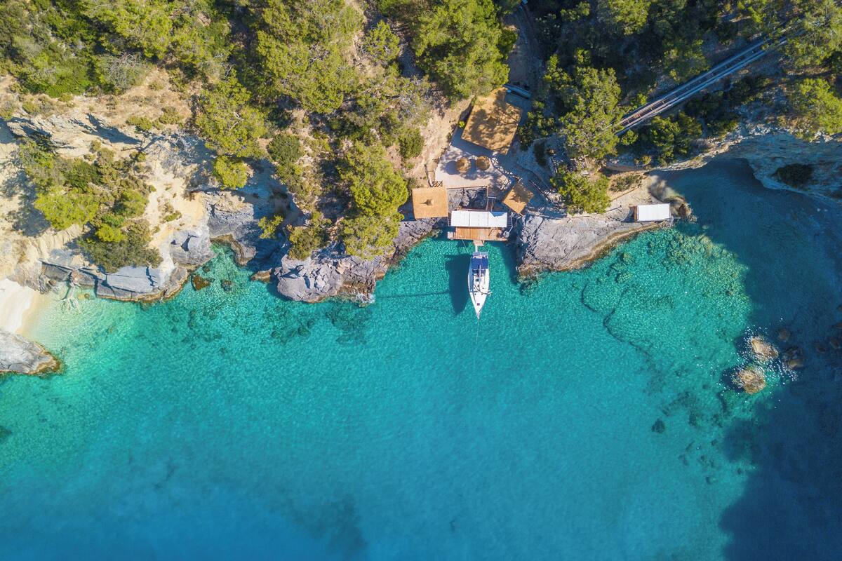 An aerial view of a coastal area with clear turquoise waters, a boat docked near a wooden structure, and rocky terrain with sparse vegetation.