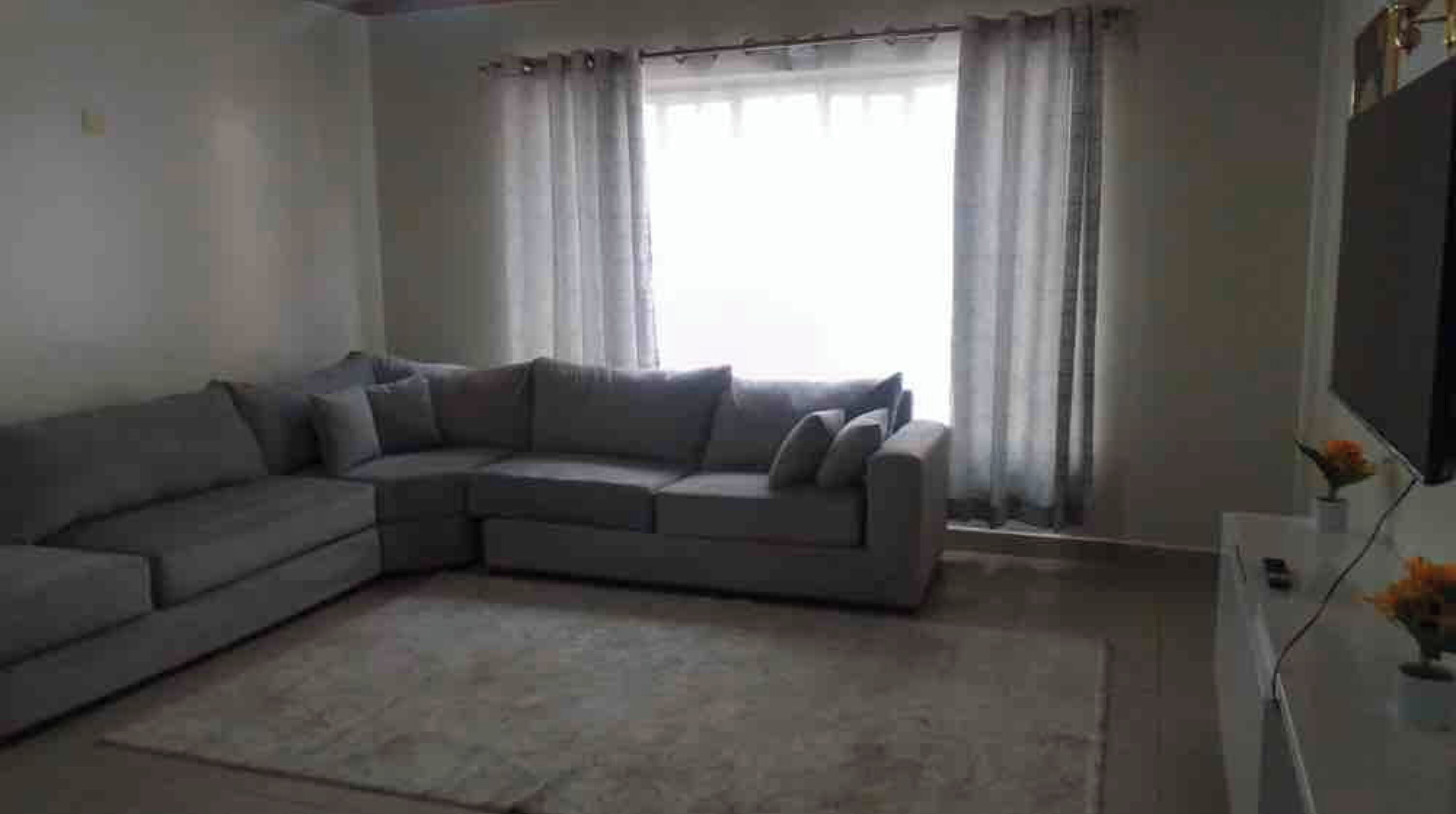 A living room with a sectional sofa, a flat-screen TV, and a large window with curtains.