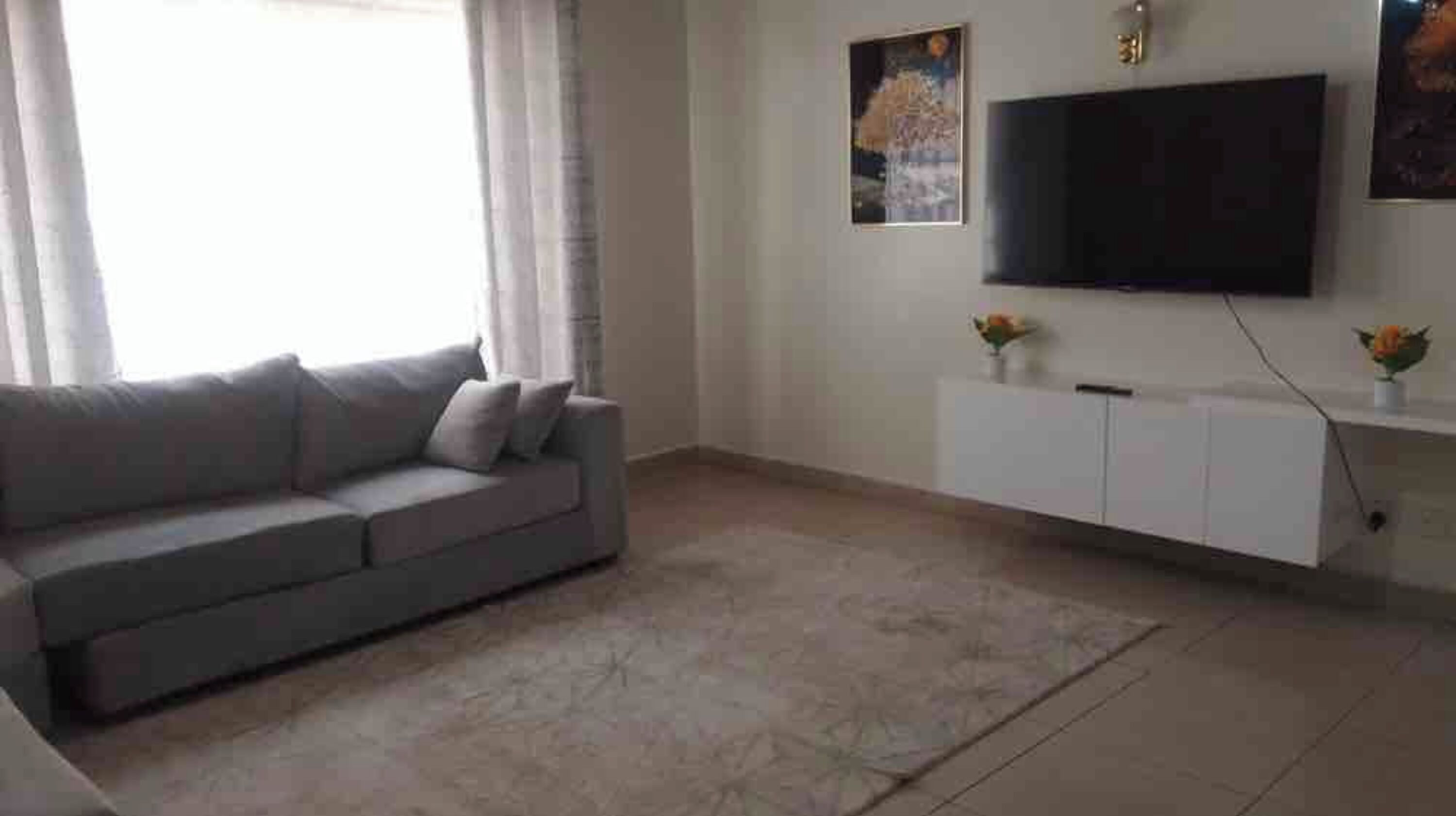 Un soggiorno con un divano grigio, una televisione a schermo piatto fissata al muro e un mobile bianco sottostante.