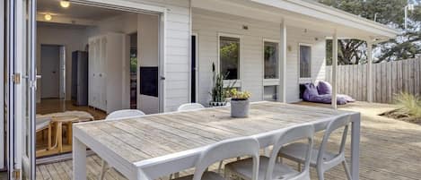 An outdoor dining area with a wooden table and white chairs, a covered patio, and a wooden fence in the background.