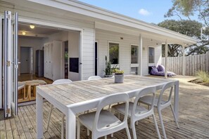 An outdoor dining area with a wooden table and white chairs, a covered patio, and a wooden fence in the background.