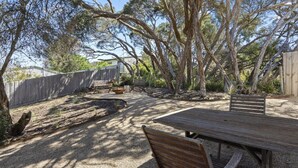 Een terras buiten met een houten tafel en stoelen, omgeven door bomen en een grindpad.